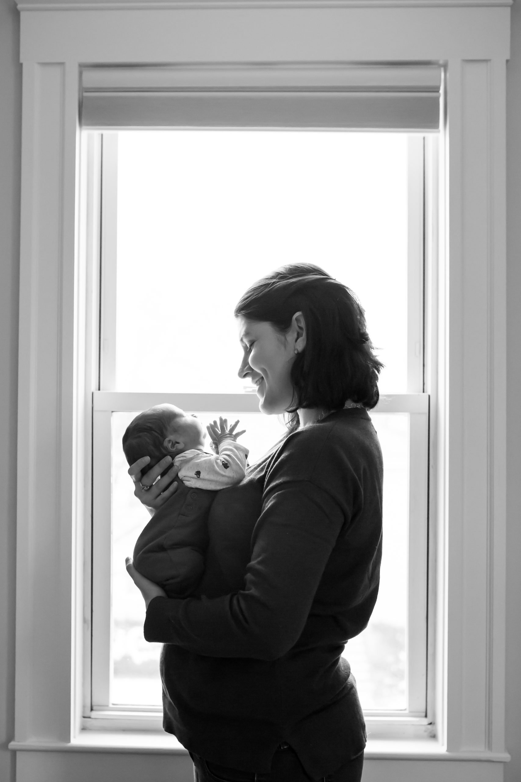 mother stands in front of window, holding her tiny newborn baby. They are looking at each other.