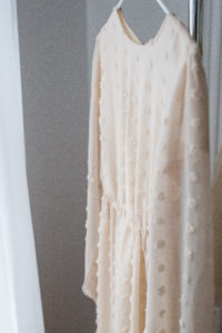 studio wardrobe: blush dotted women's dress hanging near window. long sleeves, textured dots on fabric, same color.